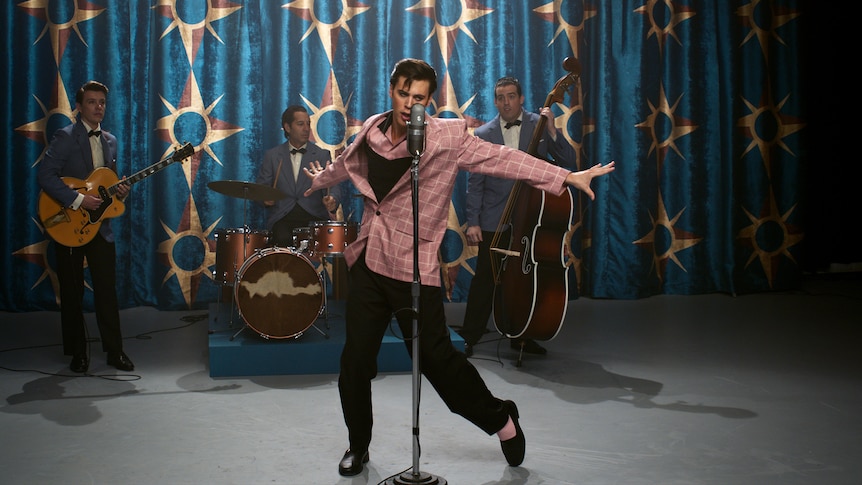 White man dressed as Elvis in a pink blazer and with quiffed hair performs on stage with a band and strikes a pose.