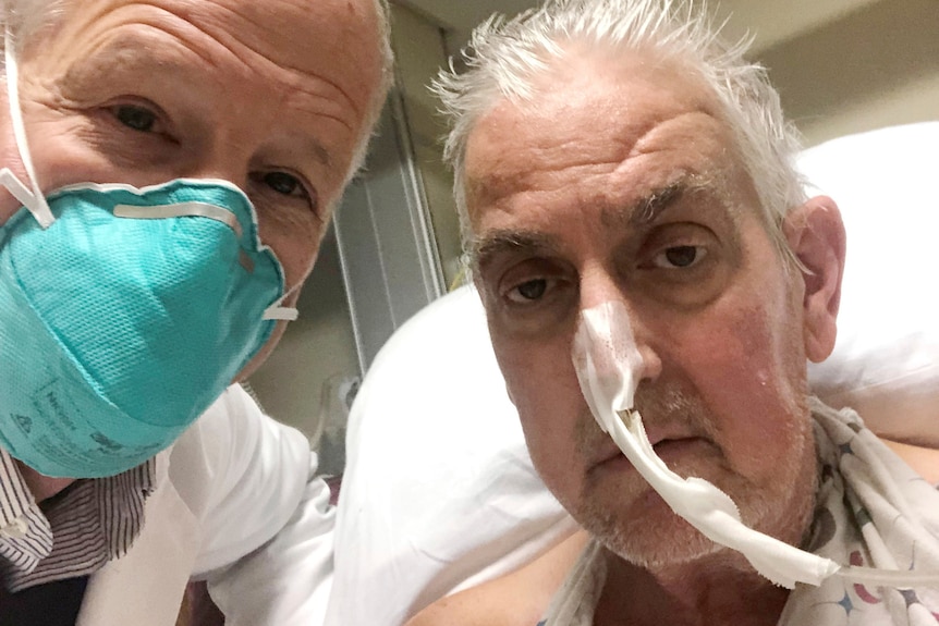Doi bărbați, unul cu un tub de oxigen în nas și celălalt purtând o mască, pozează pentru un selfie.