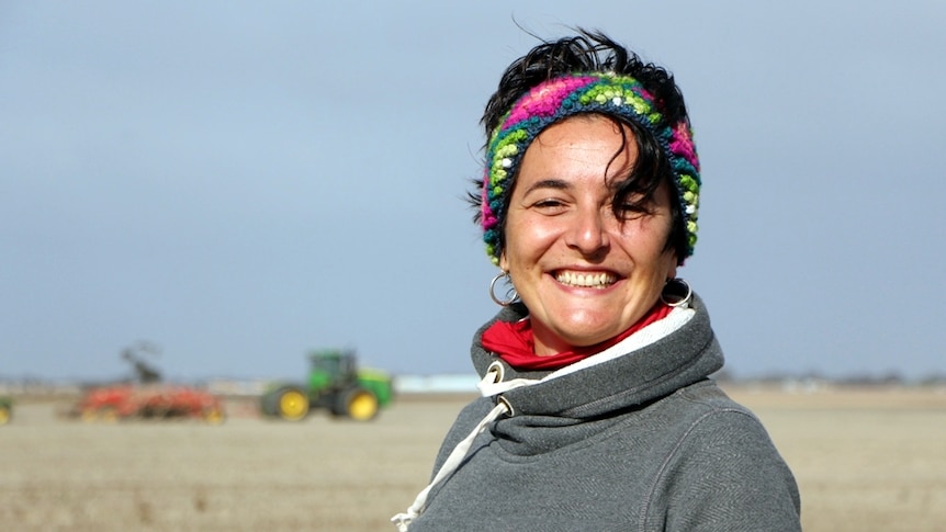 A woman standing in a paddock with a tractor sowing grain crops behind her
