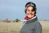 A woman standing in a paddock with a tractor sowing grain crops behind her