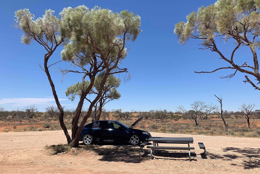 A car is seen parked next to a table and chairs with blue skies overhead, brown dirt and shrubbery behind.
