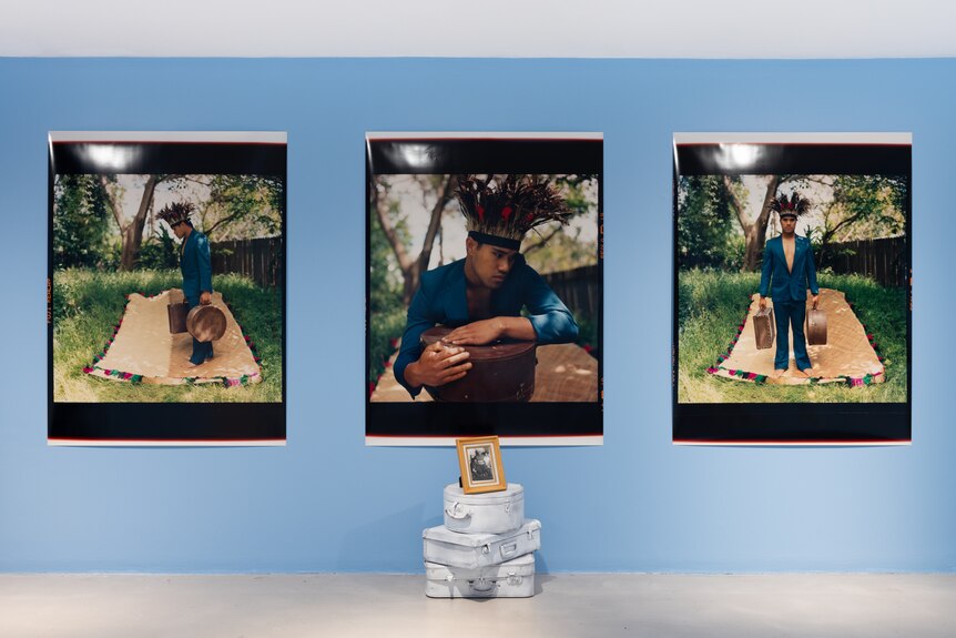 Three large art works of man in blue suit hang on blue wall. Below them, a small frame sits on suitcases.