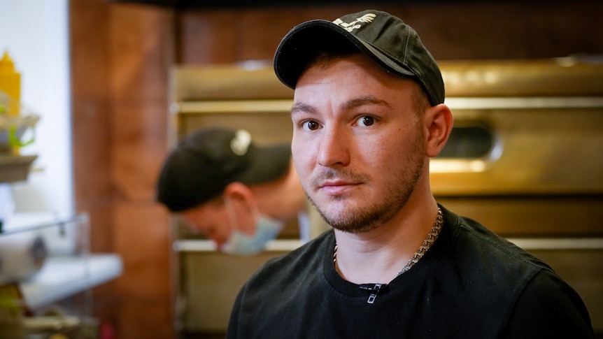 A young man in a black t-shirt and cap stands in a commercial kitchen 