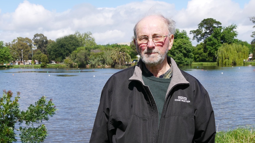 A casually dressed man with a grey beard and glasses, stands in front of a lake.