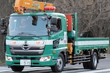 A small green truck with a flat tray back and a crane attached drives along a road in Japan.