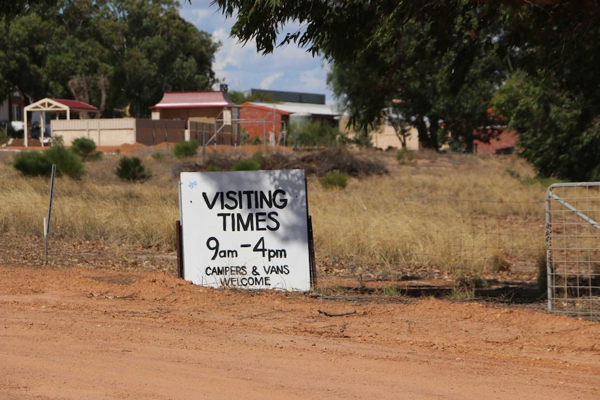 A sign on the side of a dirt road leaning up against a wire fence showing visiting times at the Principality of Hutt River.