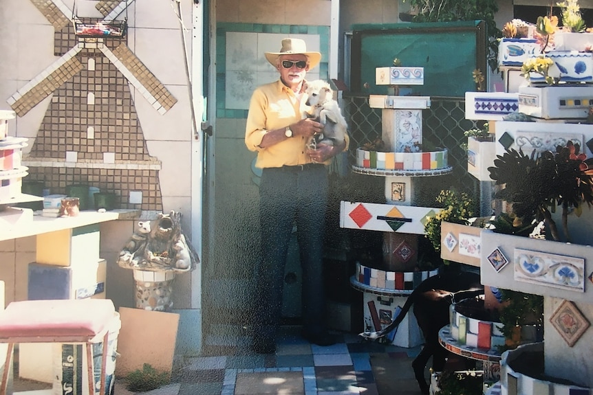 An elderly man stands in front of his tiled artworks at his house, holding his little dog.
