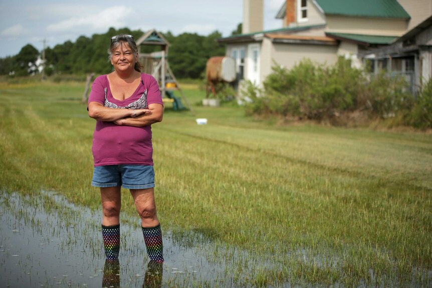 A woman in gumboots with her arms folded across her chest standing in a waterlogged front yard