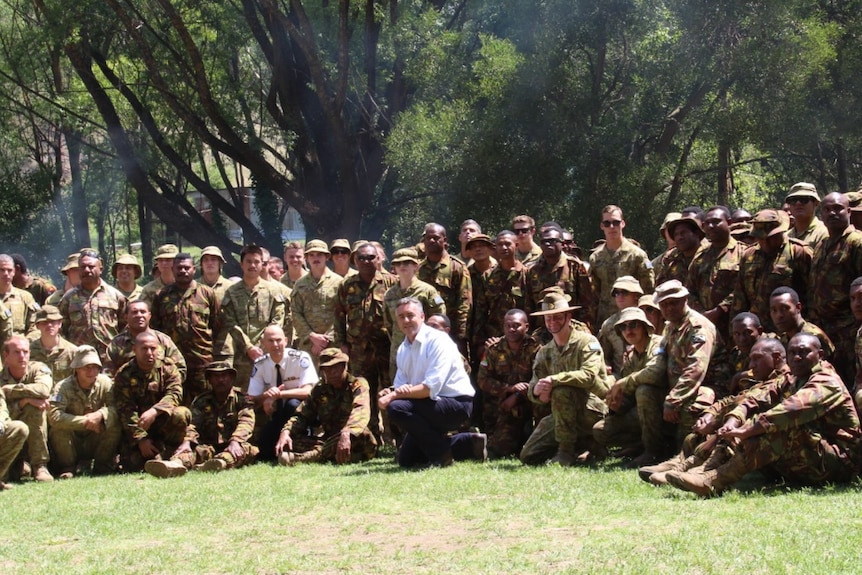 A group of people sitting under a large tree wearing army fatigues.