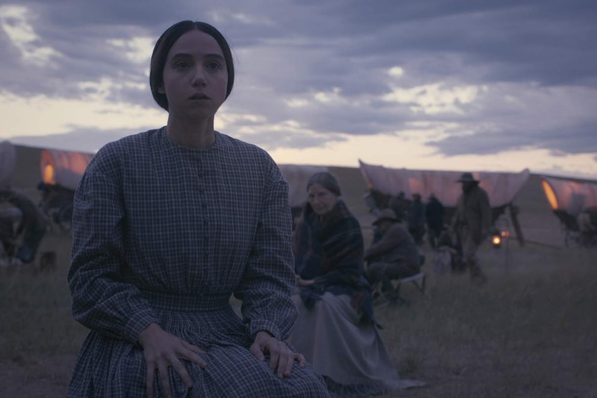 Young woman in pioneer clothing and hair in bun looks out, worried, with caravan train and twilight sky behind her.