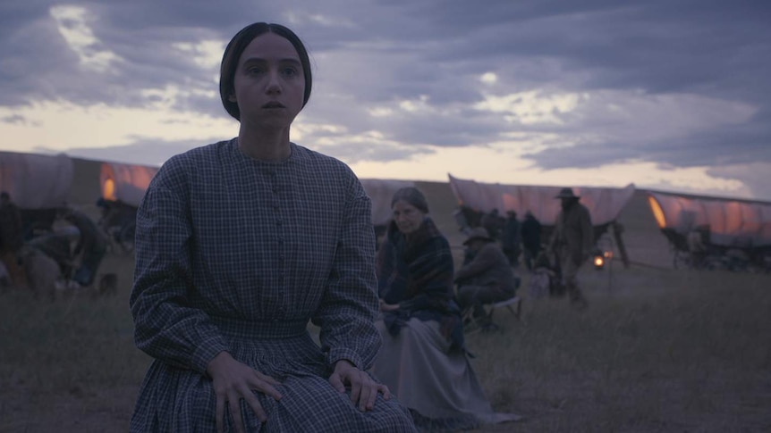 Young woman in pioneer clothing and hair in bun looks out, worried, with caravan train and twilight sky behind her.