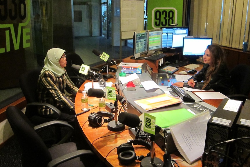 A middle-aged muslim woman in traditional garb speaks into a microphone opposite a female radio host in studio