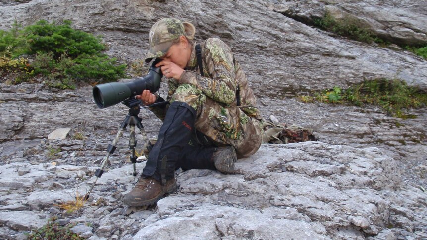 A woman in camouflage gear sitting on rocks with a telescope on a tripod.