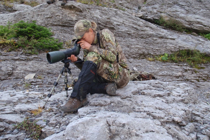 A woman in camouflage gear sitting on rocks with a telescope on a tripod.