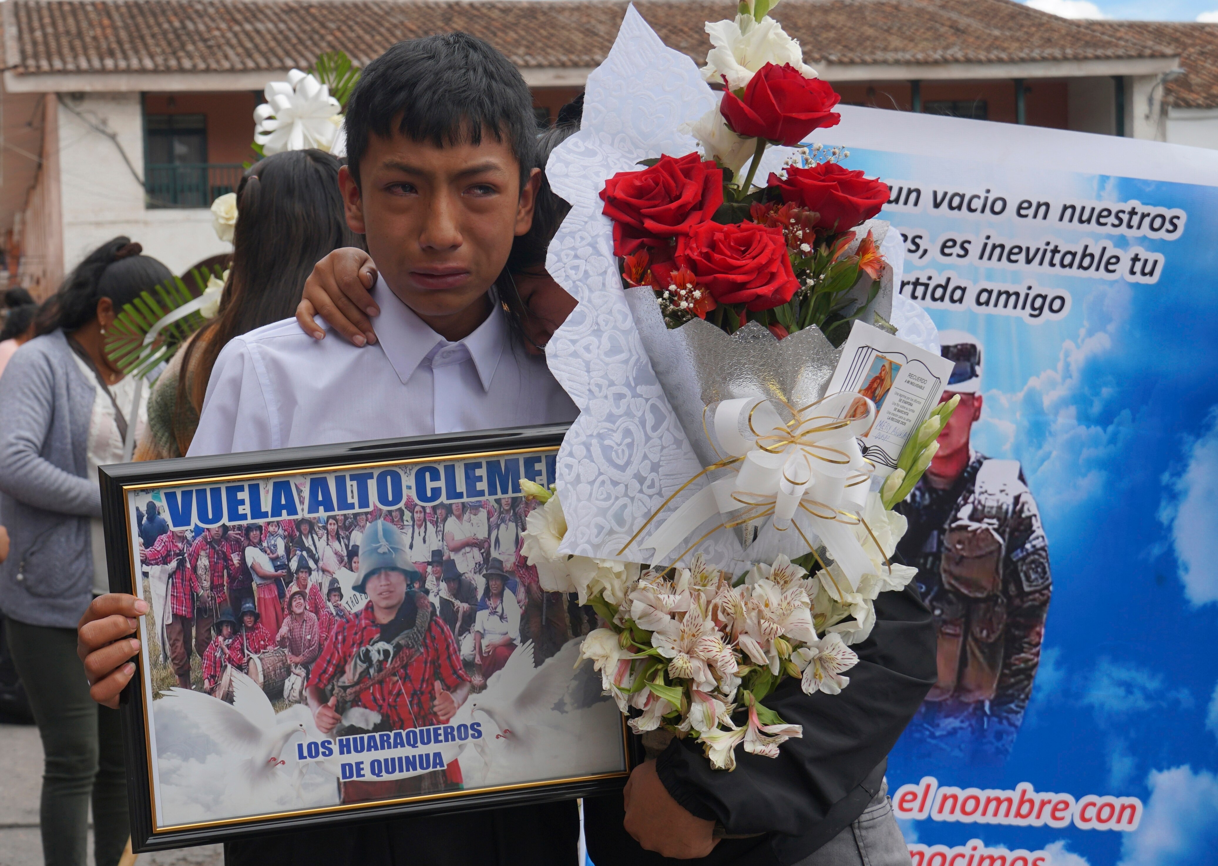 A woman holding a flower bouquet hugs a crying boy holding a frame photo. 