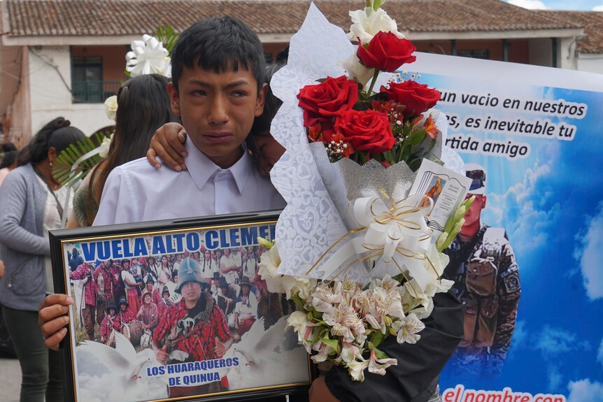 A woman holding a flower bouquet hugs a crying boy holding a frame photo. 