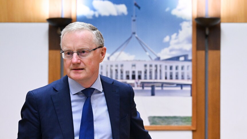 An older man wearing a blue suit and tie and glasses sits in a room with a portrait of Australia's Parliament House behind him.