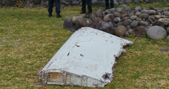 White debris from a plane sits on grass with three officials seen standing in the background, one of whom holds a radio in hand.