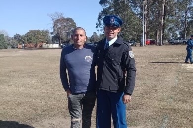An Aboriginal man wearing prison guard or police uniform stands with his arm around another man.