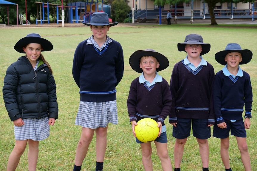 Five primary school students wearing uniform and hats stand on a football field.