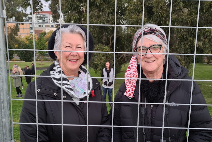 Two sisters with grey hair and black jackets smiling from behind a steel fence.