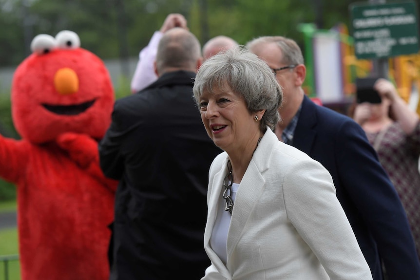 Theresa May wears a white coat and shirt, and is photobombed by an elmo impersonator at a polling booth.