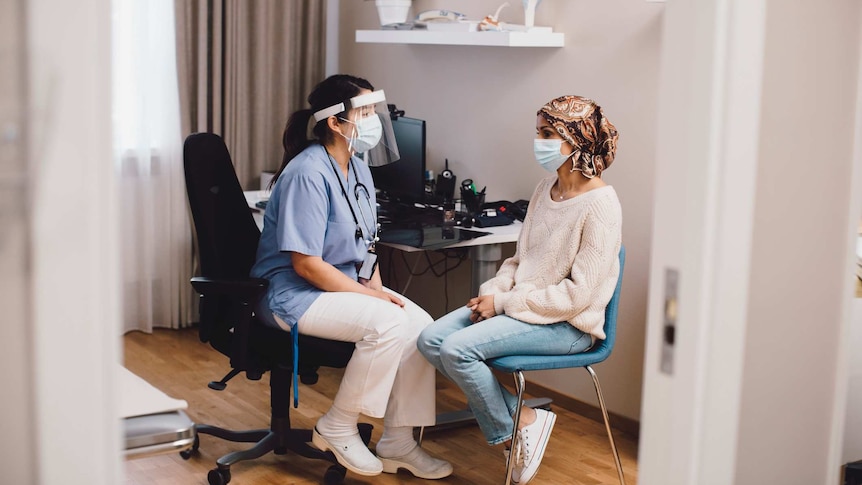 A GP wearing a face mask and extra protection talks to a patient wearing a face mask in a clinic setting
