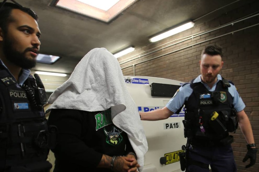 A man with a towel over his head being led by police.