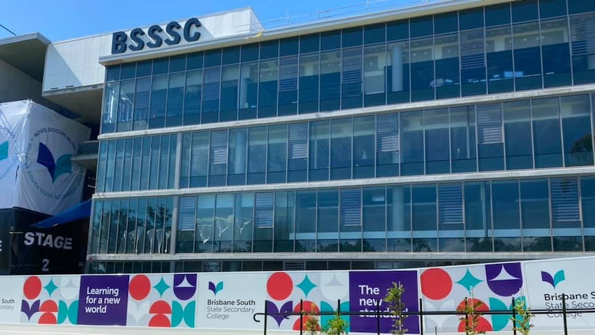 A school building with BSSSC logo in top left corner, with glass windows behind banners with school logos and grass lawn.