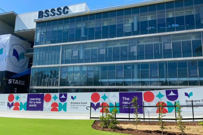 A school building with BSSSC logo in top left corner, with glass windows behind banners with school logos and grass lawn.