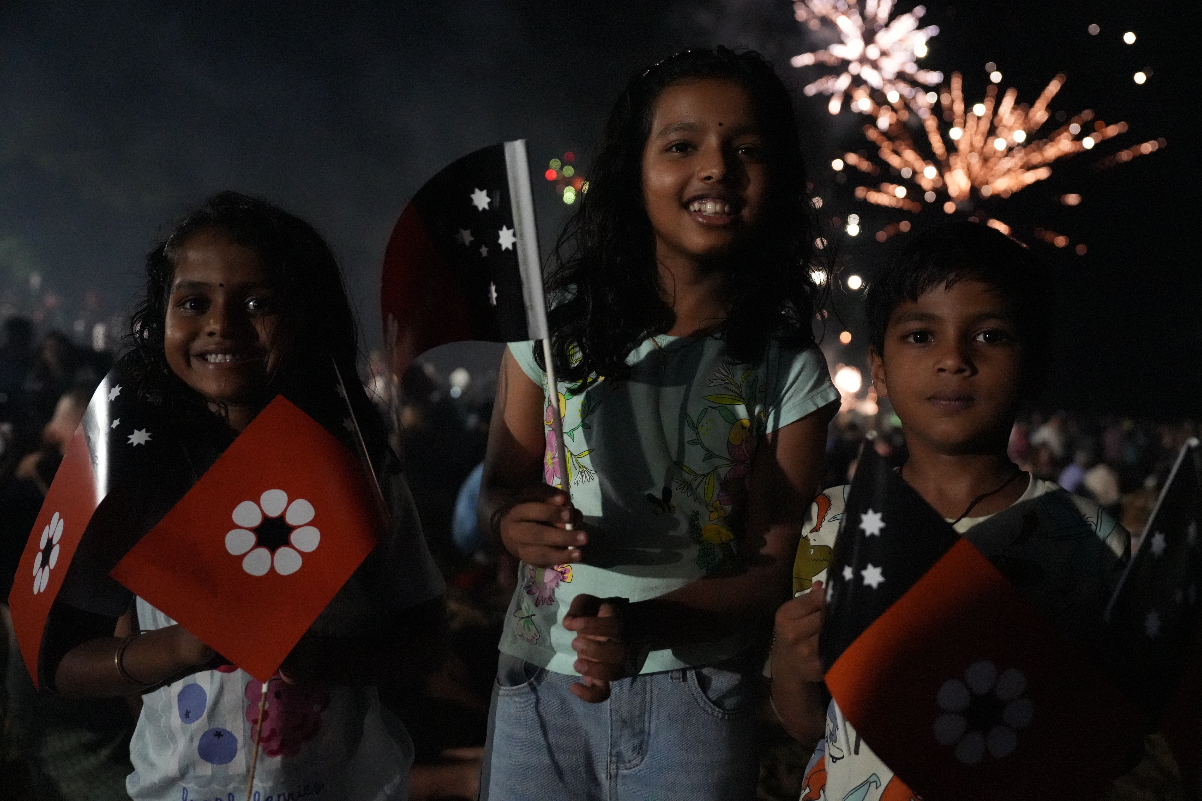 Three children wave orange flags with fireworks going off in the background
