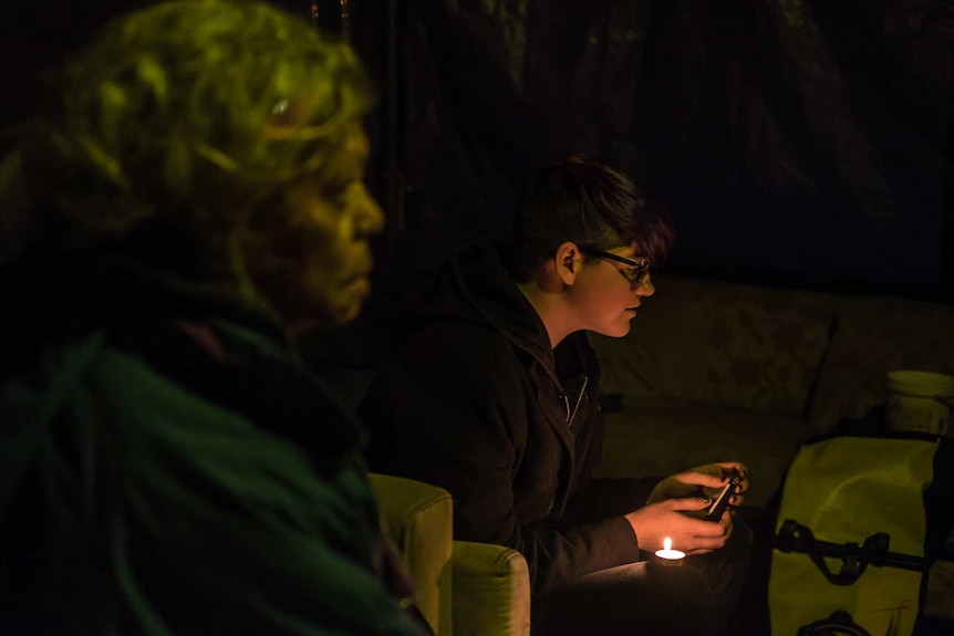 Jenny Munro and Charlie Murray sit in the 'living room' of the embassy which is lit at night by a single bulb and candles.