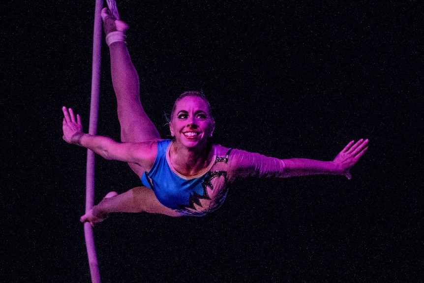 A female circus performer balances on a vertical rope, hanging on by her feet.