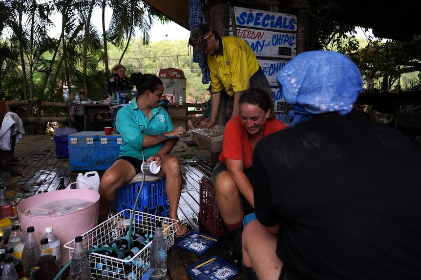 Two women and a man smile and talk to each other and they sort bottles and other items on a flood-affected deck.