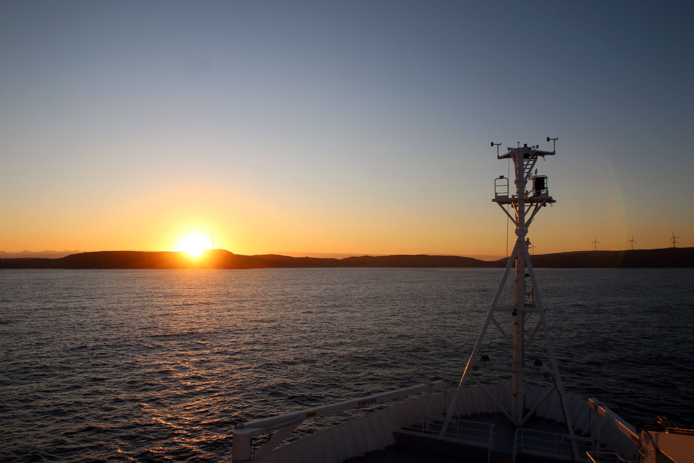 Bow of a ship with water in background and sun setting over land.