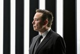 Elon Musk stands wearing a dark suit, in front of vertical black and white striped lights.
