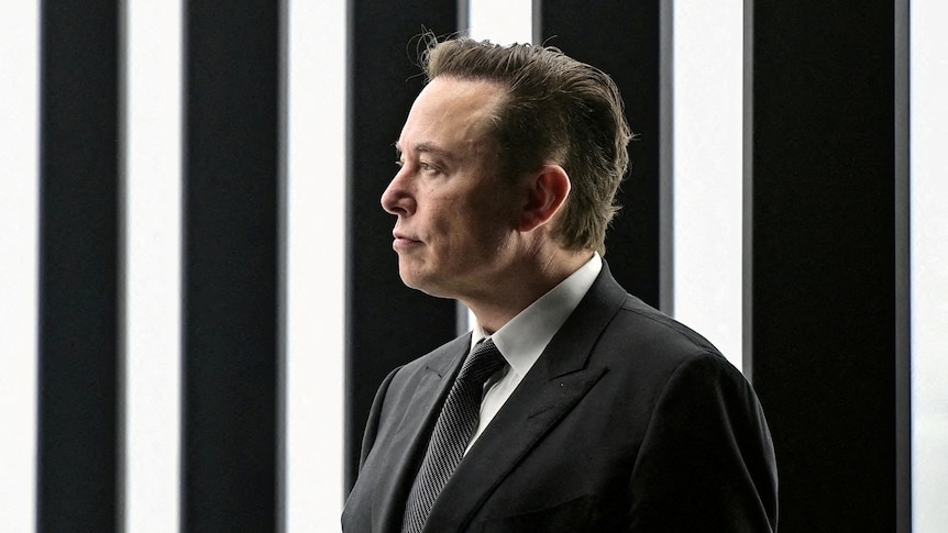 Elon Musk stands wearing a dark suit, in front of vertical black and white striped lights.