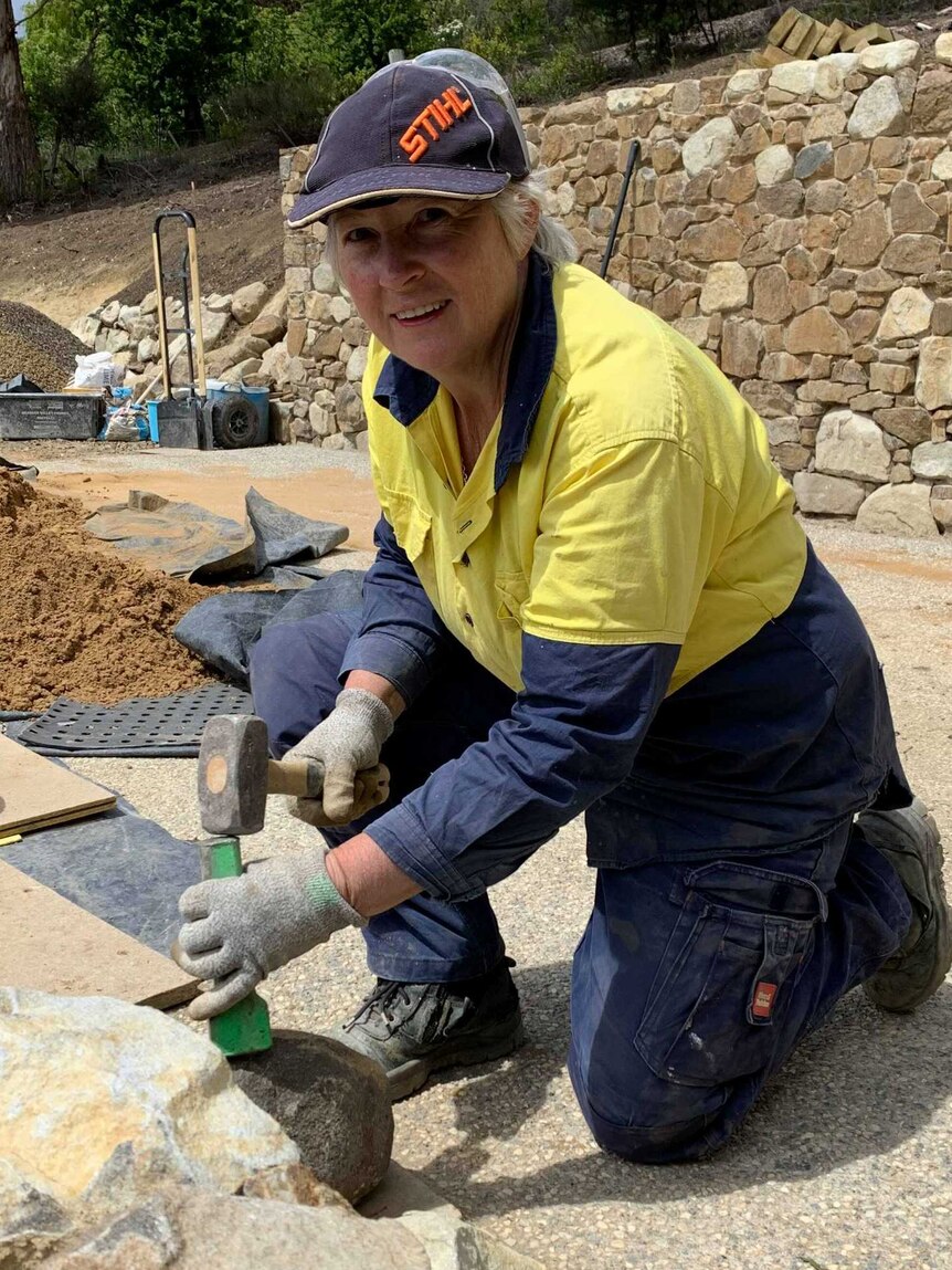 Lyn Stedman hitting stones with a hammer, with a stone wall in the background, Tasmania 2019.