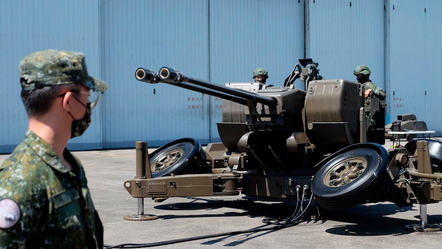 A man in a Taiwanese military uniform watches as soldiers operate a large, double-barrelled caon on an open concrete space.