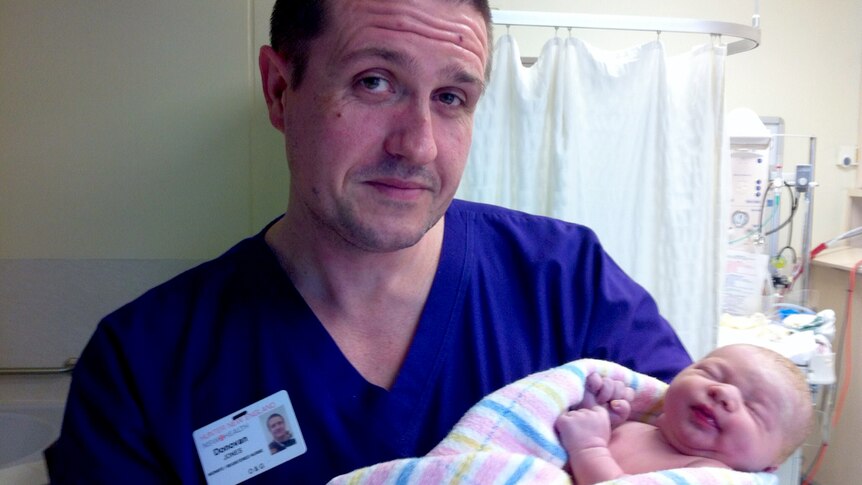 A man in blue hospital scrubs holding a new-born baby.