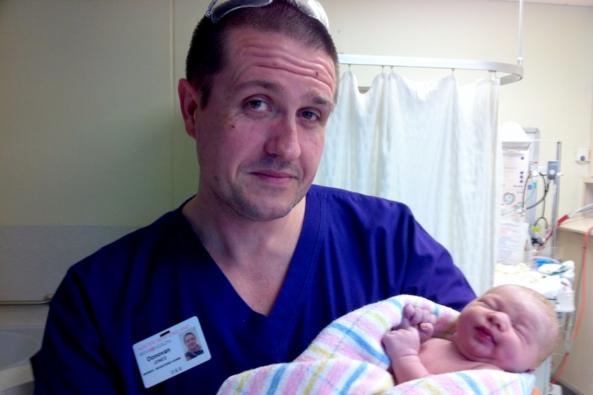 A man in blue hospital scrubs holding a new-born baby.