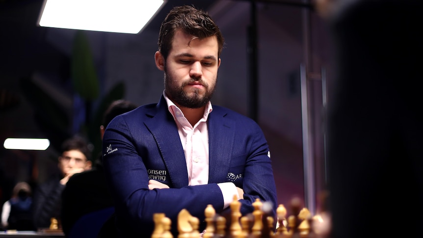 A chess grandmaster in a blue jacket sits with his arms folded looking down at the pieces on the board.