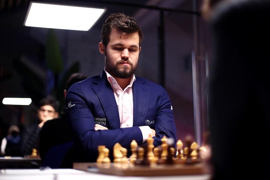 A chess grandmaster in a blue jacket sits with his arms folded looking down at the pieces on the board.