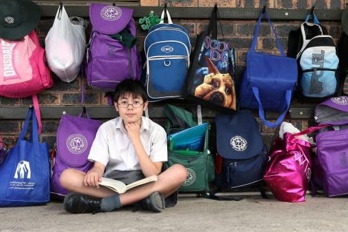 A teenage boy sitting on the ground in front of a school bag rack.