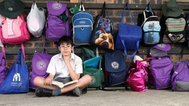 A teenage boy sitting on the ground in front of a school bag rack.