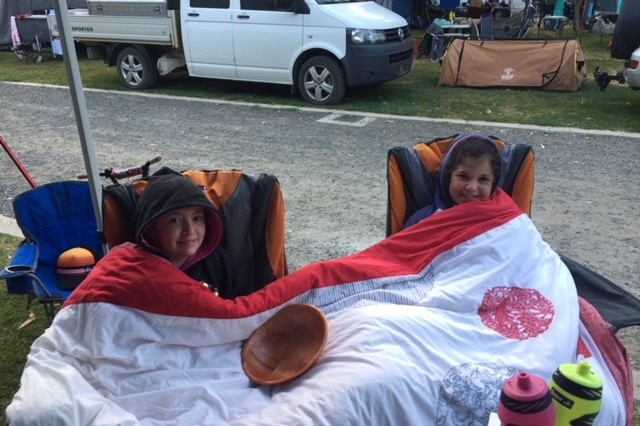 Two kids in camping chairs, resting under a quilt, at a caravan park.