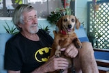 Bill, older man with grey hair and facial hair wearing Batman shirt and holding small brown dog smiling.