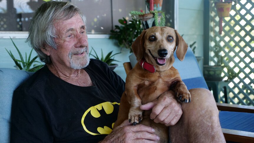 Bill, older man with grey hair and facial hair wearing Batman shirt and holding small brown dog smiling.