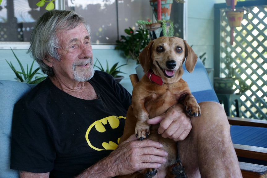 Bill, older man with grey hair and facial hair wearing batman shirt and holding small brown dog smiling.