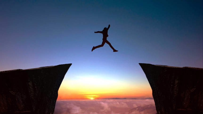 Person in silhouette leaping between two cliff edges. Clouds and sunset or sunrise in the background.